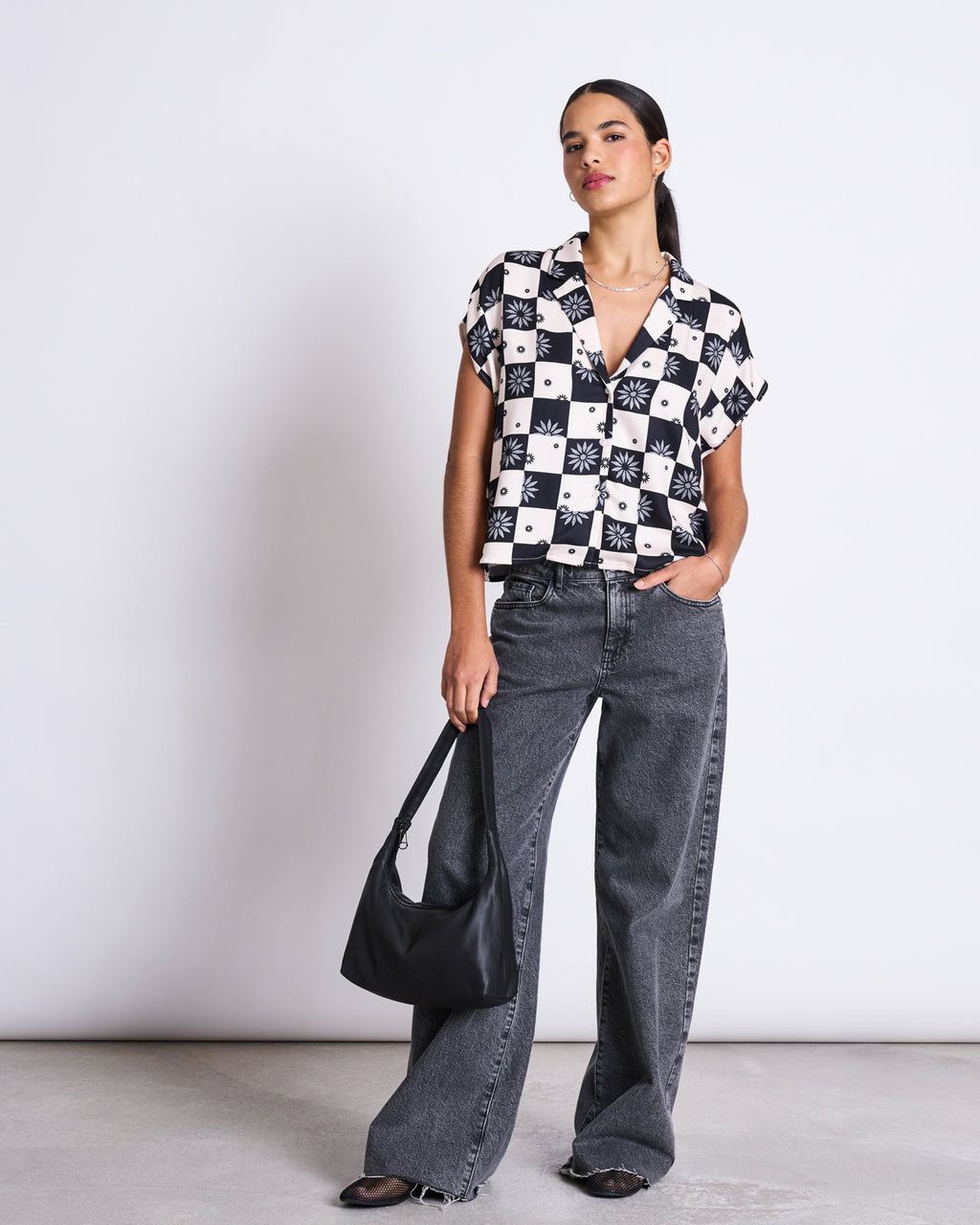 A woman stands against a plain background wearing the jan-n-june BLOUSE TIERRA FLORAL SQUARE PRINT, paired with loose gray jeans, brown shoes, and holding a black shoulder bag in her left hand.