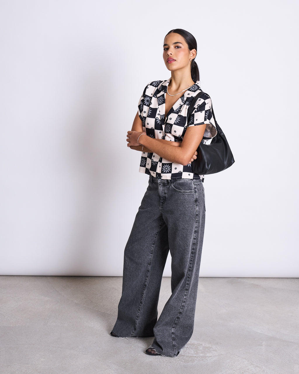 A woman stands against a white wall wearing the jan-n-june BLOUSE TIERRA FLORAL SQUARE PRINT, loose gray jeans, black sandals, and carrying a black shoulder bag. She looks slightly to the side with her arms crossed.