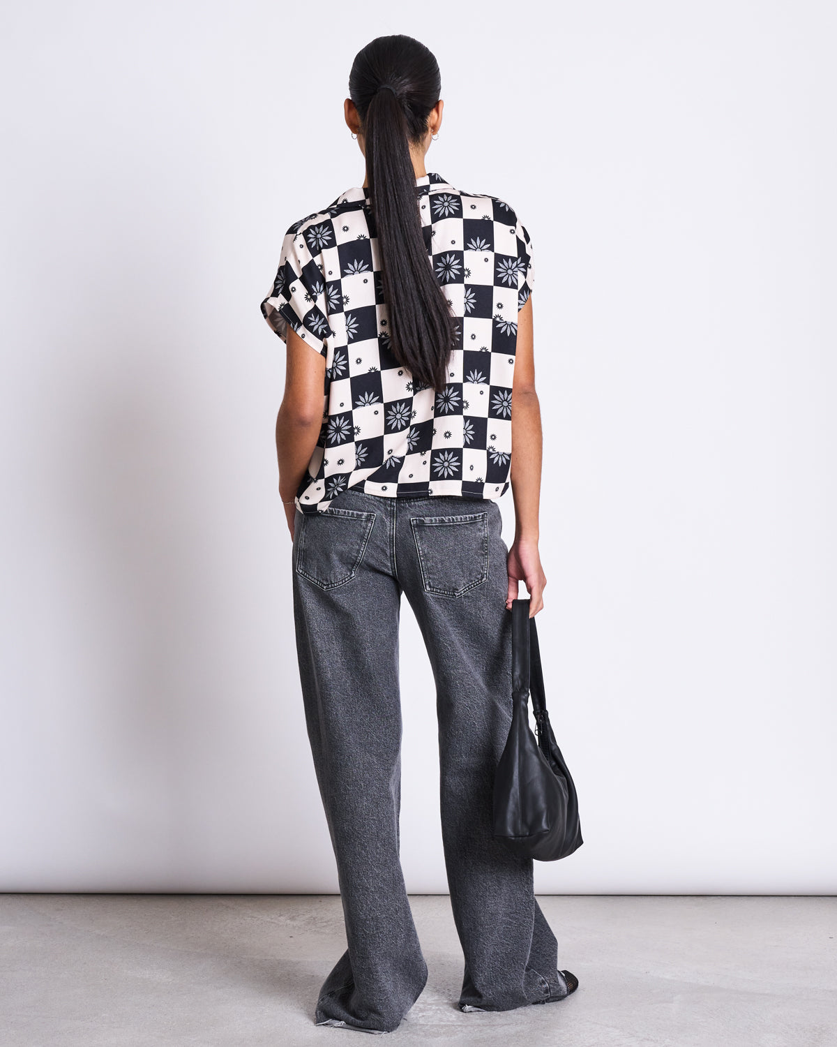A person with long dark hair in a low ponytail faces away, wearing the jan-n-june BLOUSE TIERRA FLORAL SQUARE PRINT, loose gray jeans, and holding a black bag by their side, standing against a plain background.