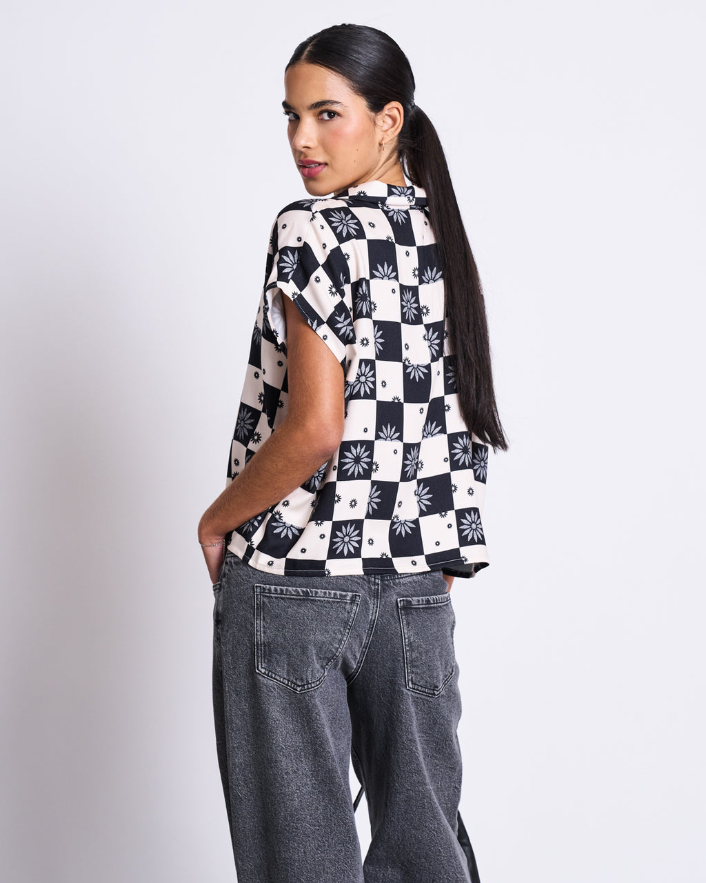 A woman with long dark hair in a ponytail wears the jan-n-june BLOUSE TIERRA FLORAL SQUARE PRINT with black-and-white geometric and floral patterns, paired with loose gray jeans, standing against a plain white background.