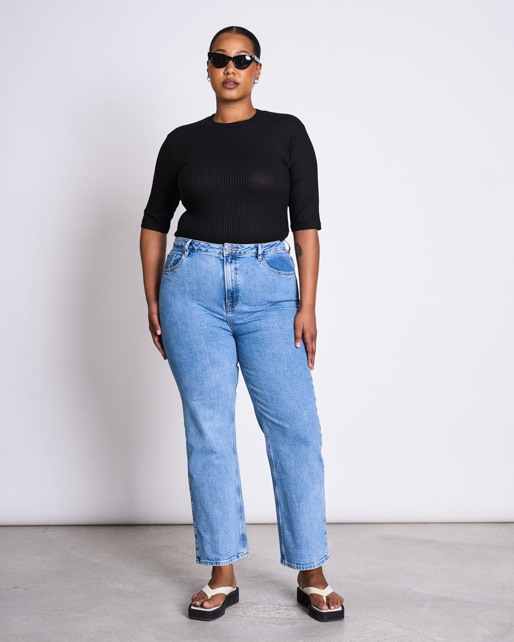 A woman wearing jan-n-junes LYOCELL RIB T-SHIRT NINA BLACK, black sunglasses, light blue high-waisted jeans, and black platform sandals stands against a plain white wall.