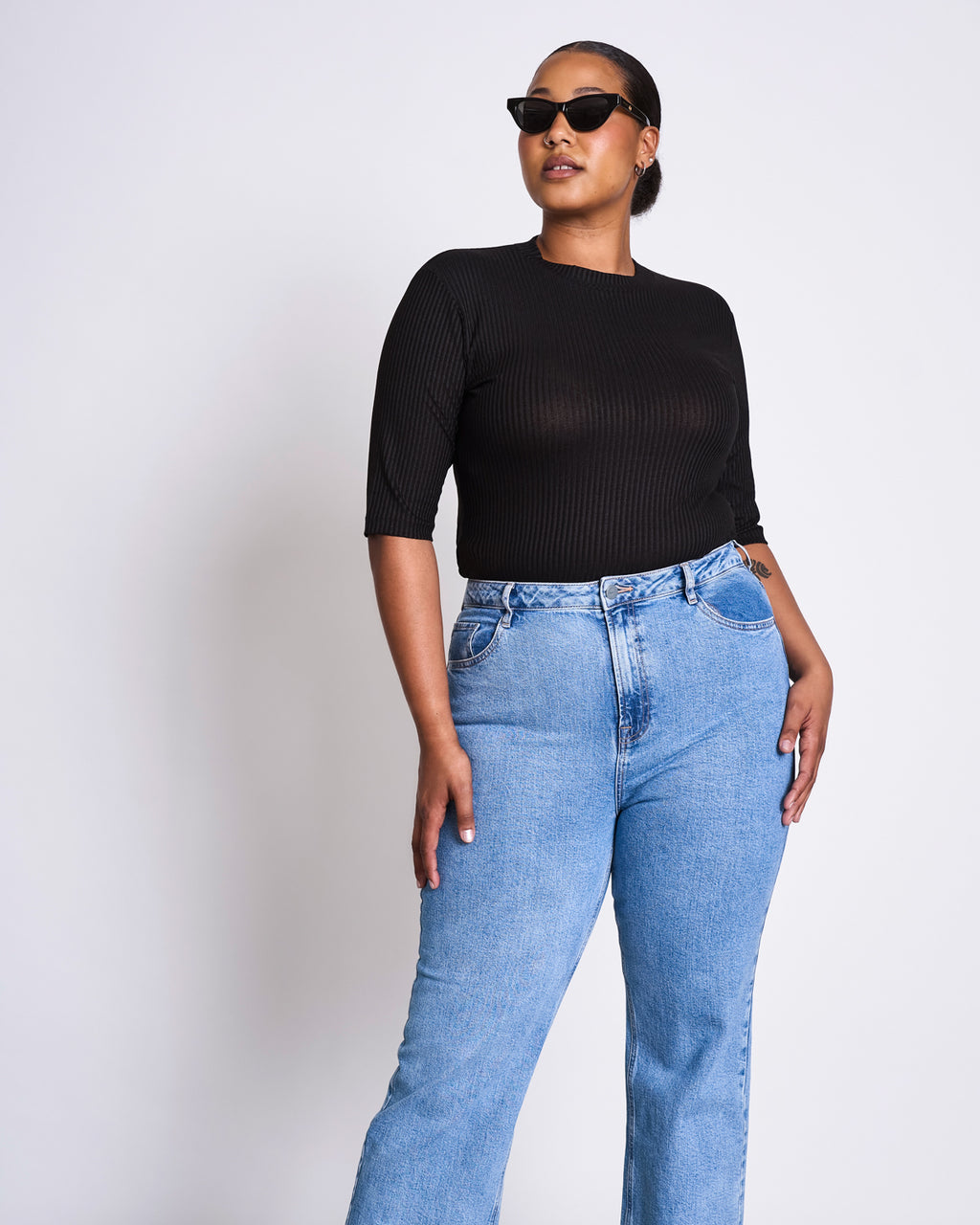 A woman in cat-eye sunglasses, the jan-n-june LYOCELL RIB T-SHIRT NINA BLACK, and high-waisted blue jeans poses confidently against a plain white background.