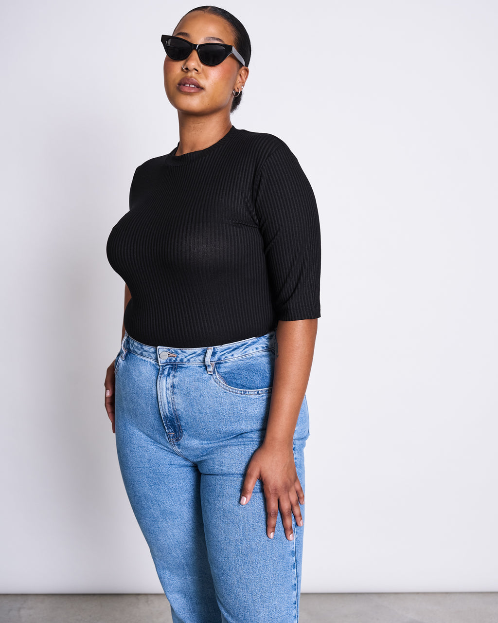 A woman in cat-eye sunglasses, the jan-n-june LYOCELL RIB T-SHIRT NINA BLACK with a mini turtleneck and elbow-length sleeves, and high-waisted blue jeans stands confidently against a plain white background.