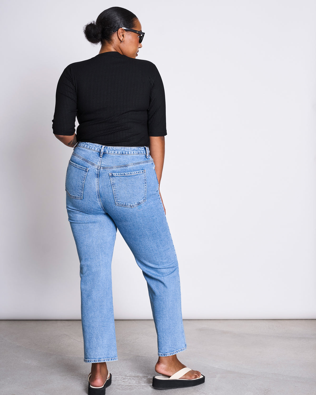 A woman faces away, wearing the jan-n-june LYOCELL RIB T-SHIRT NINA BLACK, light blue high-waisted jeans, black sunglasses, and black-and-white slide sandals against a plain light background.