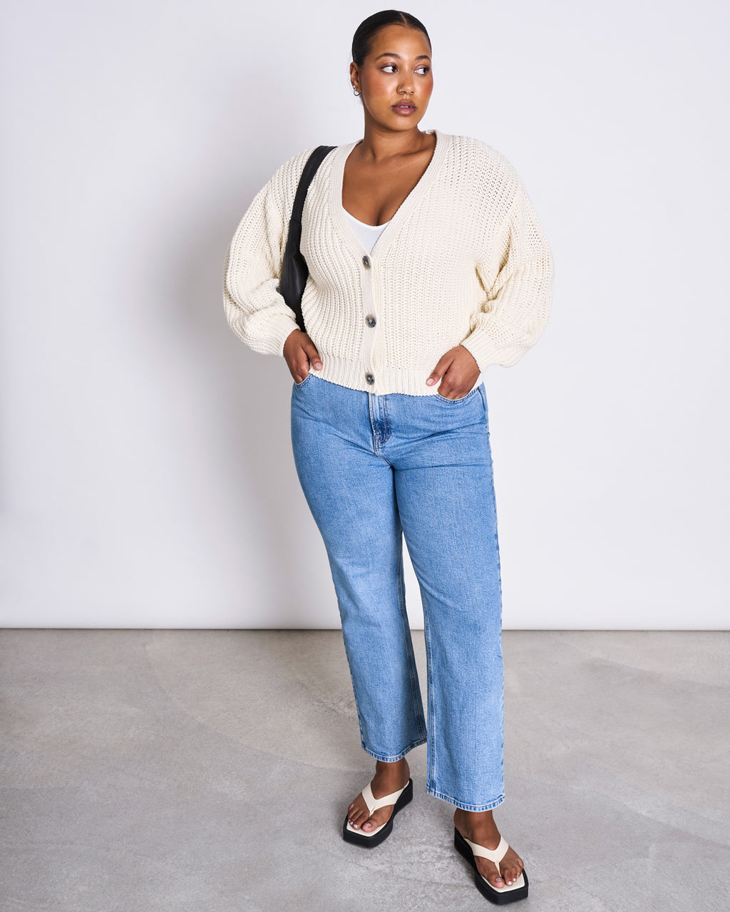 A woman stands against a white wall wearing the jan-n-june CHUNKY CARDIGAN LENA OFFWHITE GOTS over a white top, blue jeans, and black sandals, carrying a black shoulder bag. Her hair is pulled back and her hands are in her cardigan pockets.