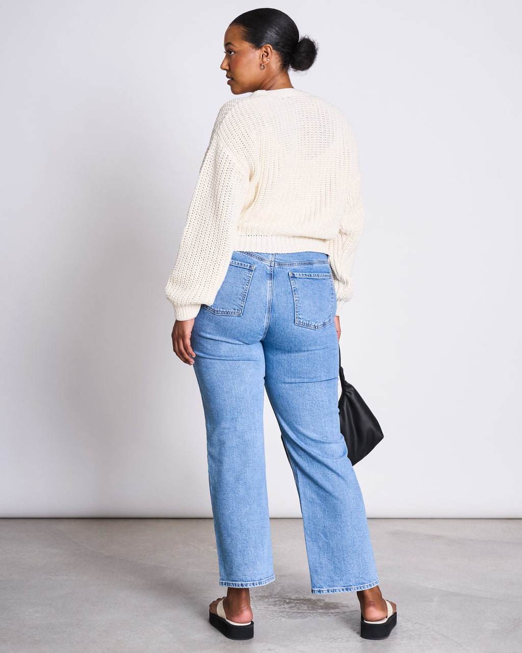 A woman with her hair in a neat bun stands with her back to the camera, wearing the jan-n-june CHUNKY CARDIGAN LENA OFFWHITE GOTS, light blue high-waisted jeans, black sandals, and holding a small black bag.