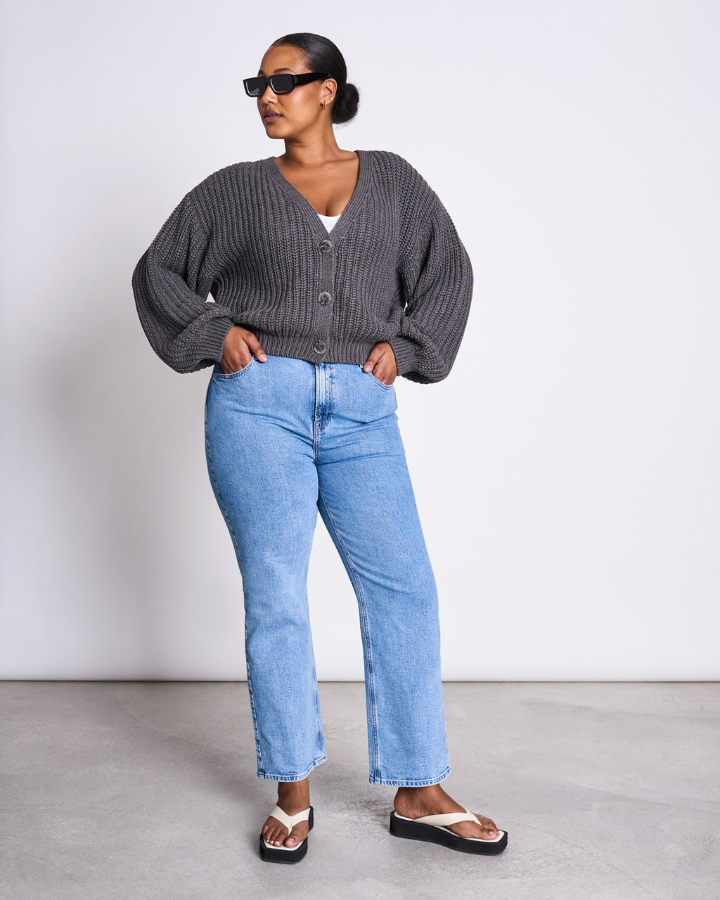 A woman wearing sunglasses, light blue jeans, flip-flop sandals, and the jan-n-june CHUNKY CARDIGAN LENA DARK GREY GOTS stands against a plain white wall with her hands in her pockets.