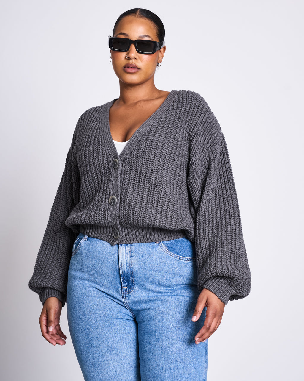 A woman stands against a plain white background, wearing black sunglasses, high-waisted blue jeans, and the jan-n-june CHUNKY CARDIGAN LENA DARK GREY GOTS over a white top.