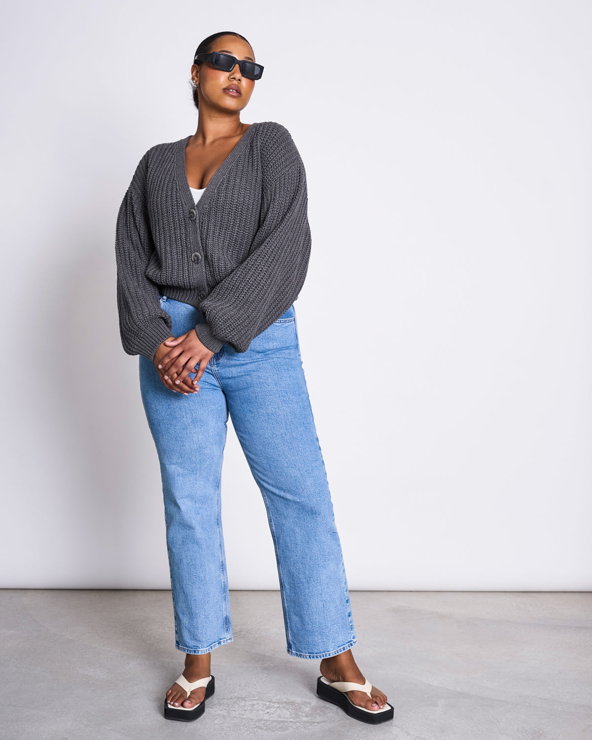 A woman wearing sunglasses, the jan-n-june CHUNKY CARDIGAN LENA DARK GREY GOTS over a white top, light blue jeans, and black sandals stands against a plain white wall with her hands clasped in front of her.