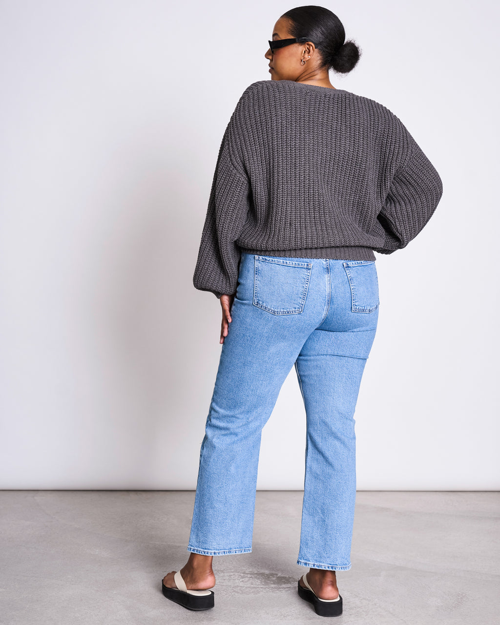 A person with dark hair in a bun wears the jan-n-june CHUNKY CARDIGAN LENA DARK GREY GOTS, light blue jeans, black sunglasses, and black sandals. They stand indoors in a minimal setting, facing away from the camera.