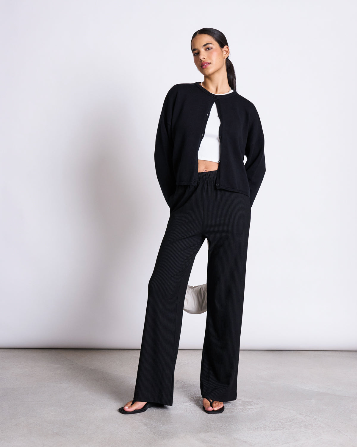A woman poses against a white wall wearing the jan-n-june LIGHT CARDIGAN ASAS BLACK GOTS over a white organic cotton crop top, paired with black wide-leg pants and sandals. Her dark hair is pulled back and one hand rests behind her hip.