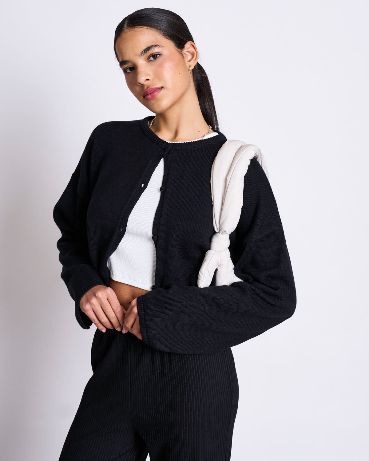 A woman poses against a plain background wearing the jan-n-june LIGHT CARDIGAN ASAS BLACK GOTS over a white crop top, black ribbed pants, and carries a padded off-white bag over her shoulder.