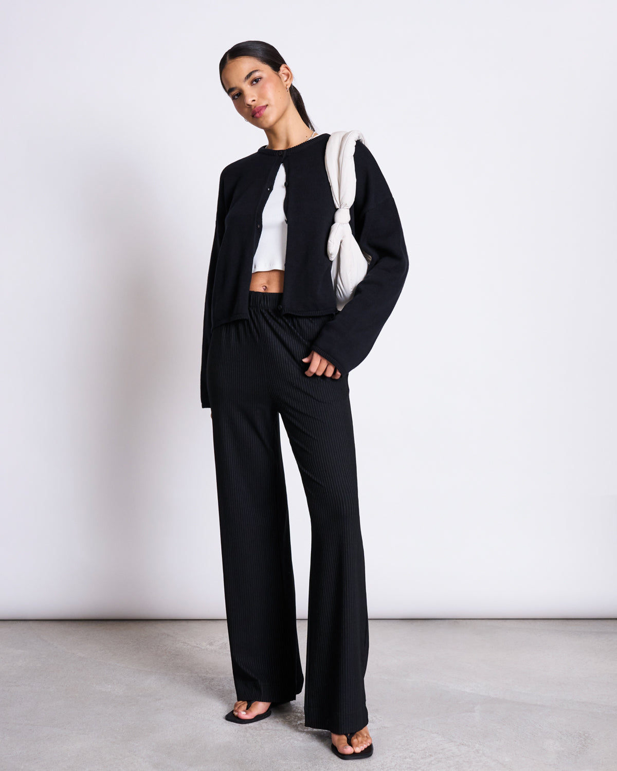 A woman poses against a white wall wearing the jan-n-june LIGHT CARDIGAN ASAS BLACK GOTS, black wide-leg pants, a white organic cotton crop top, black heeled sandals, and a cream sweater draped over her shoulder.