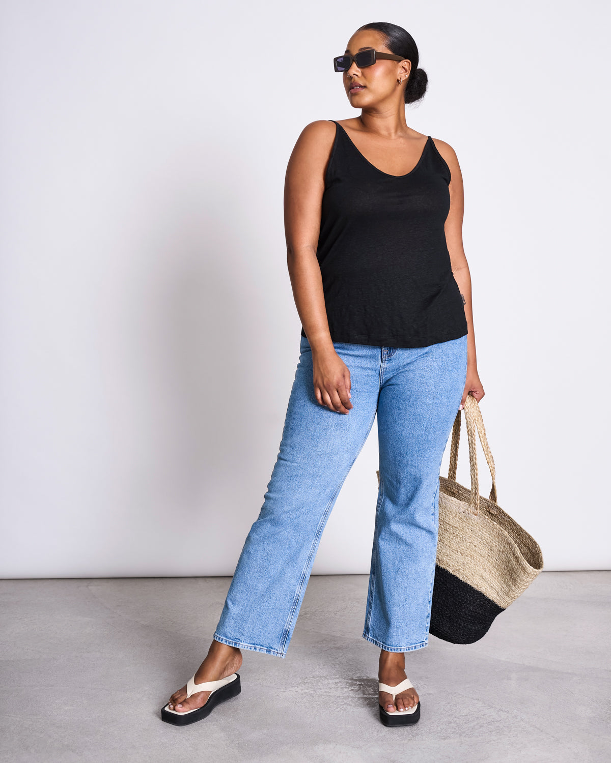 A woman wearing sunglasses, a black sleeveless top, light blue jeans, and white sandals stands against a plain background holding the jan-n-june SHOPPING BASKET JUTE BLACK.