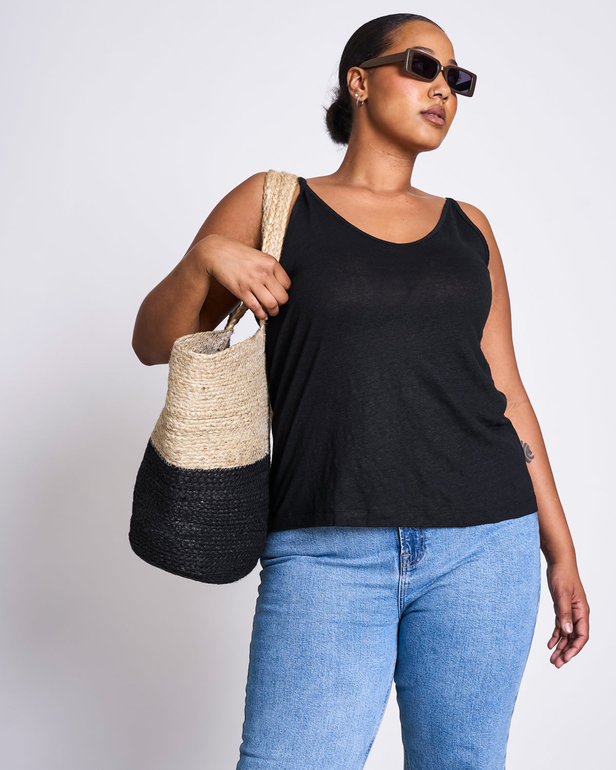 A woman stands confidently against a white background, wearing sunglasses and holding the jan-n-june SHOPPING BASKET JUTE BLACK over her shoulder, showcasing modern style and conscious living.