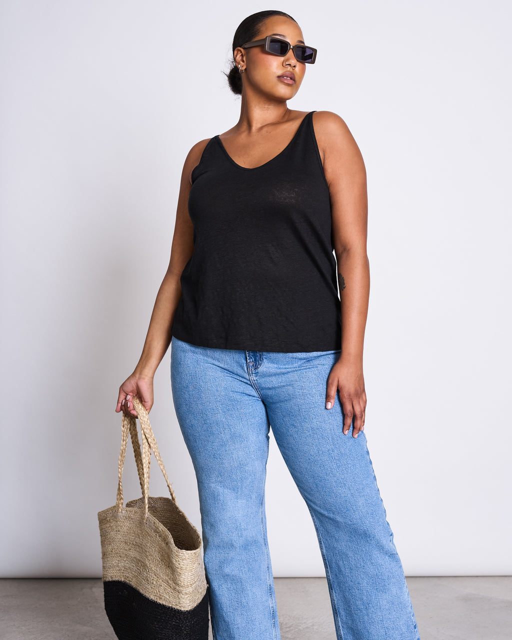 A woman wearing sunglasses, a black sleeveless top, and light blue jeans confidently poses with the jan-n-june SHOPPING BASKET JUTE BLACK against a plain white background.