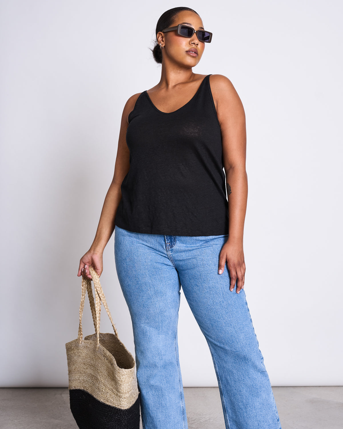 A woman wearing sunglasses, a black sleeveless top, and light blue jeans confidently poses with the jan-n-june SHOPPING BASKET JUTE BLACK against a plain white background.
