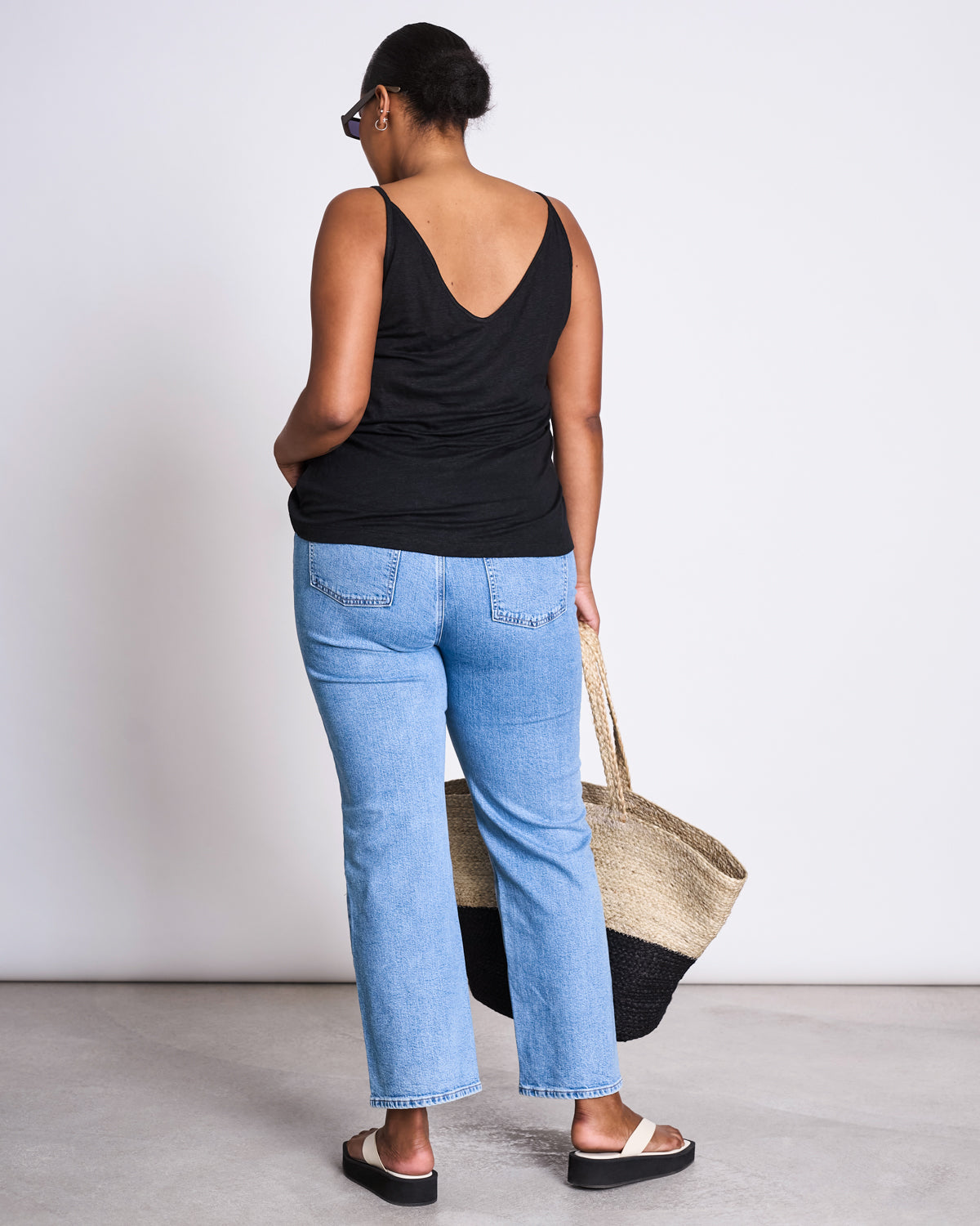A woman faces away in a plain indoor space, wearing a black sleeveless top, light blue jeans, black-and-white sandals, sunglasses, and holding the jan-n-june SHOPPING BASKET JUTE BLACK.