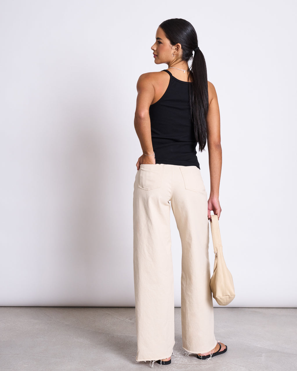 A woman with long dark hair in a ponytail stands with her back to the camera against a white background, wearing the jan-n-june RIB TOP STEM BLACK, beige wide-leg pants, black sandals, and holding a beige handbag.
