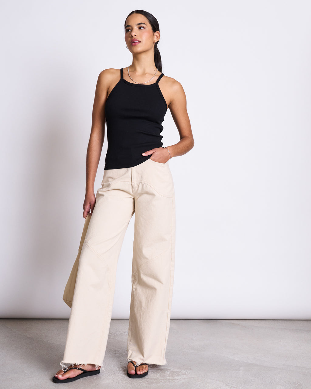 A woman stands before a white wall, wearing the jan-n-june RIB TOP STEM BLACK with ribbed texture, paired with light beige wide-leg pants and black sandals. Her hair is pulled back, one hand in her pocket, gazing slightly to the side.