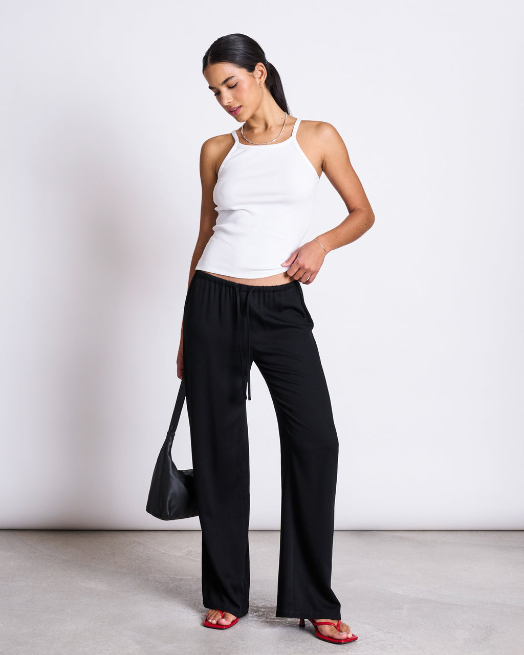 A woman stands against a white background wearing the jan-n-june RIB TOP STEM WHITE, black wide-leg pants, red sandals, and a black shoulder bag. She looks down and touches her pants pocket with one hand.