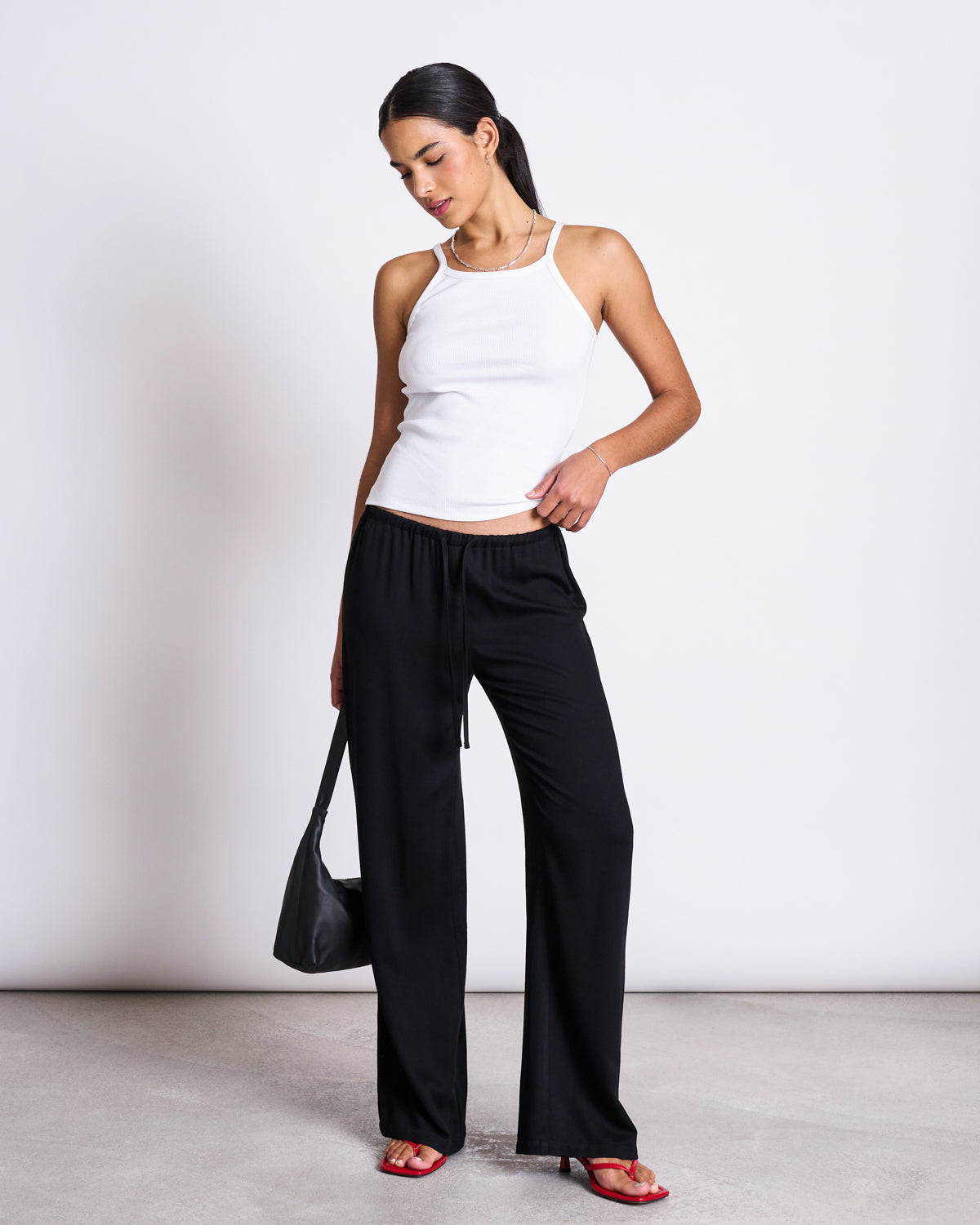 A woman stands against a white background wearing the jan-n-june RIB TOP STEM WHITE, black wide-leg pants, red sandals, and a black shoulder bag. She looks down and touches her pants pocket with one hand.