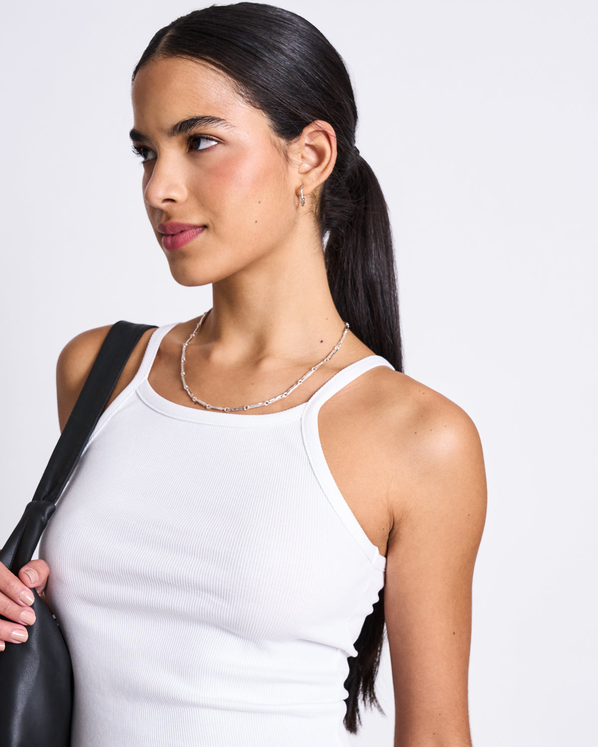 A woman with long dark hair in a low ponytail wears the jan-n-june RIB TOP STEM WHITE, styled with a silver necklace, small hoop earrings, and a black shoulder bag. She looks slightly to the side against a plain background.