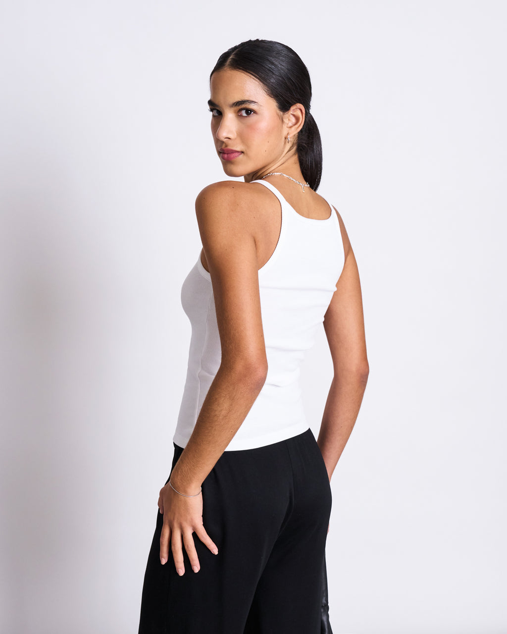 A woman with dark hair in a low ponytail wears the jan-n-june RIB TOP STEM WHITE and black pants, standing sideways and looking over her shoulder against a plain white background.