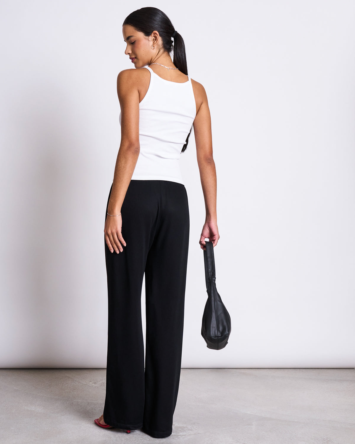 A woman with long dark hair in a ponytail models the jan-n-june RIB TOP STEM WHITE, paired with black wide-leg pants and holding a black handbag, standing against a plain white background.