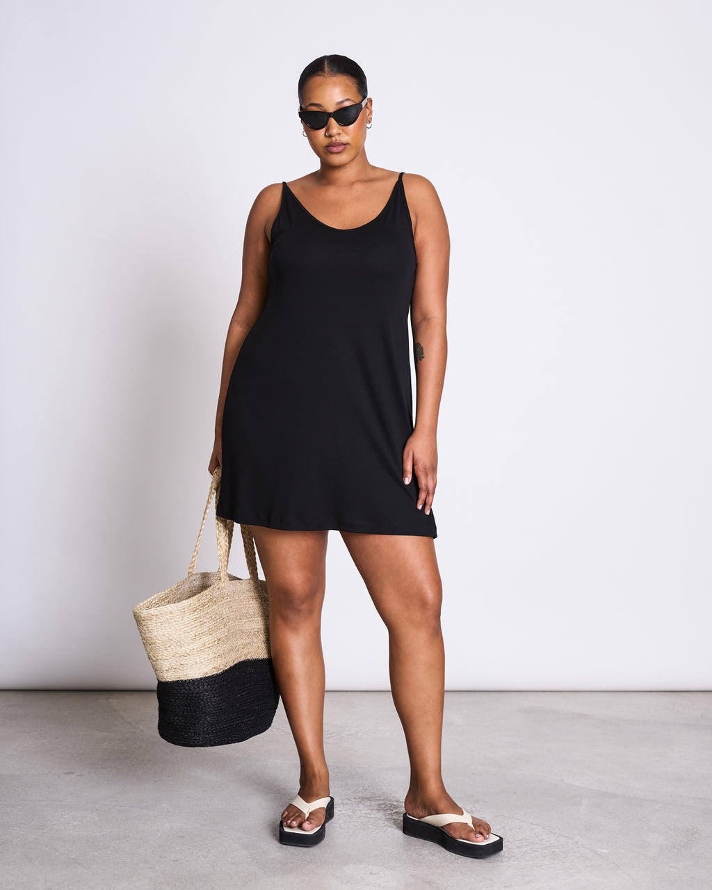 A woman stands confidently in jan-n-junes MINI DRESS TRIANGLE BLACK with black sunglasses, sandals, and a woven straw bag with black trim—capturing effortless summer style against a plain white background.