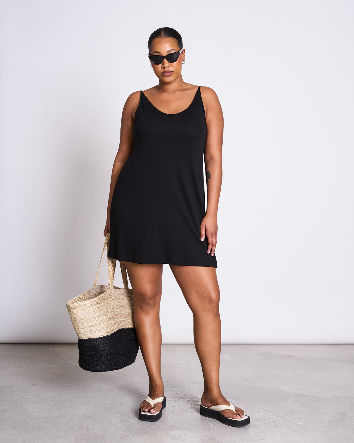 A woman stands confidently in jan-n-junes MINI DRESS TRIANGLE BLACK with black sunglasses, sandals, and a woven straw bag with black trim—capturing effortless summer style against a plain white background.