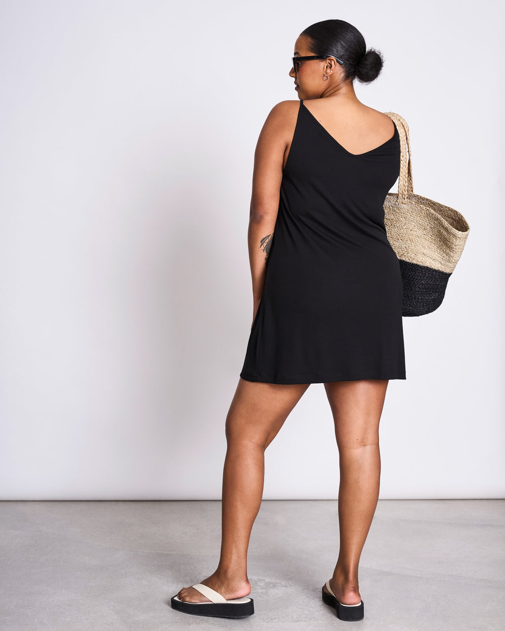 A woman faces away, wearing jan-n-junes MINI DRESS TRIANGLE BLACK—a sleeveless sustainable slip dress—with black sandals, sunglasses, and a woven tote bag over her shoulder against a plain light background.