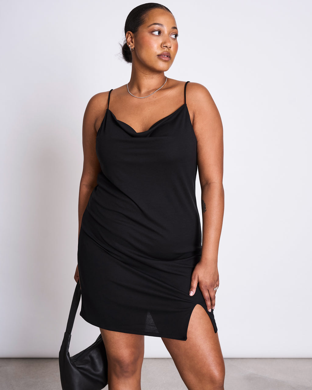 A woman wearing the jan-n-june MINI DRESS HELEN BLACK with a waterfall neckline stands against a white background, holding a black handbag and looking to the side.