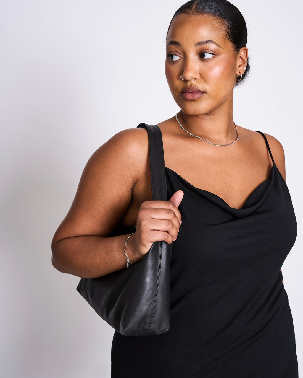 A woman with a sleek low bun wears the jan-n-june MINI DRESS HELEN BLACK and carries a black shoulder bag, gazing to her right against a plain white background.