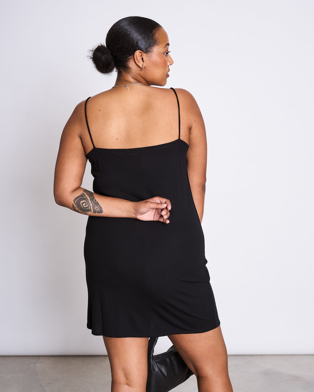 A woman with a tattoo on her left upper arm and her hair in a low bun stands facing away, one hand behind her back, wearing the jan-n-june MINI DRESS HELEN BLACK—a mini length dress with thin straps.