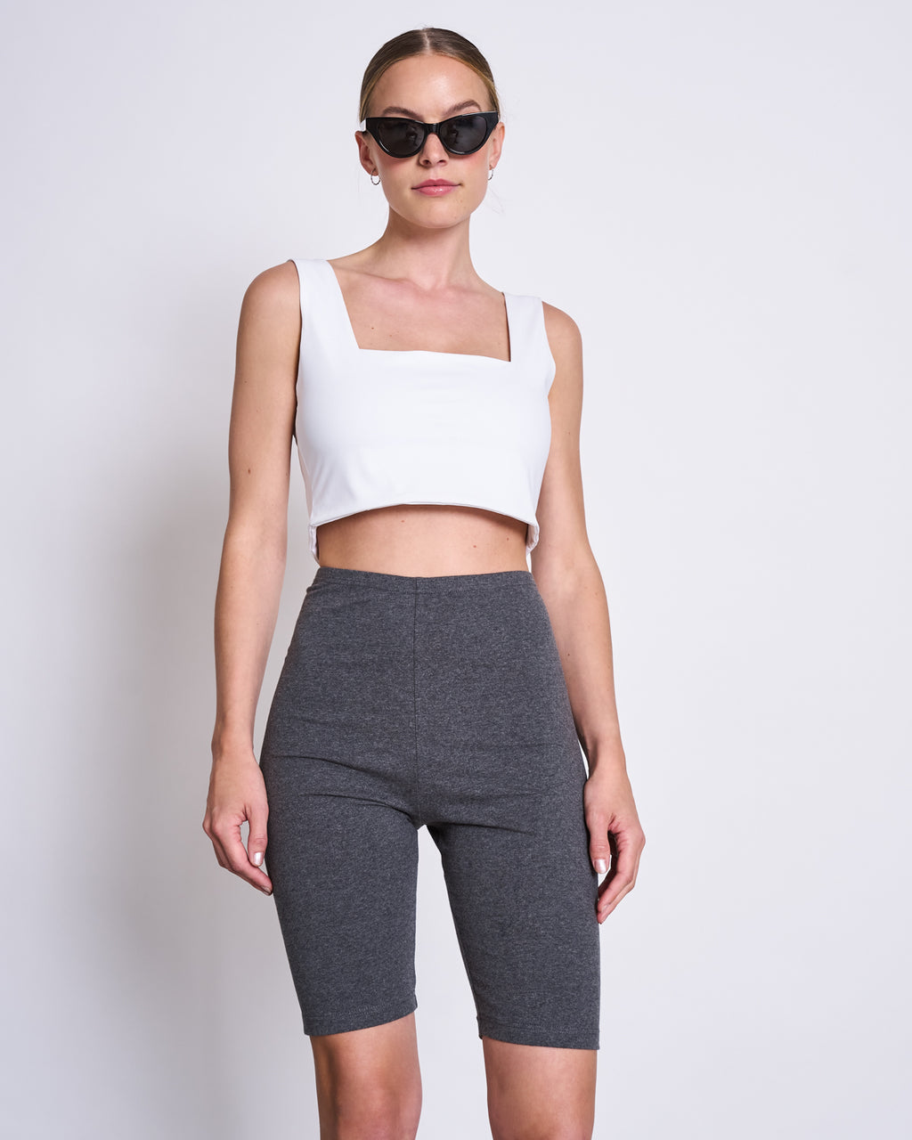A woman stands against a plain white background wearing jan-n-junes CROP TOP CANCUN WHITE, a white sleeveless crop top with a square neckline and neoprene-like fabric, along with black sunglasses and high-waisted gray biker shorts.