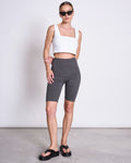 A woman in jan-n-june BIKER SHORTS DARK GREY, a white square-neck crop top, black platform sandals, and sunglasses poses against a plain light background, radiating effortless style with an Angel Skin glow.