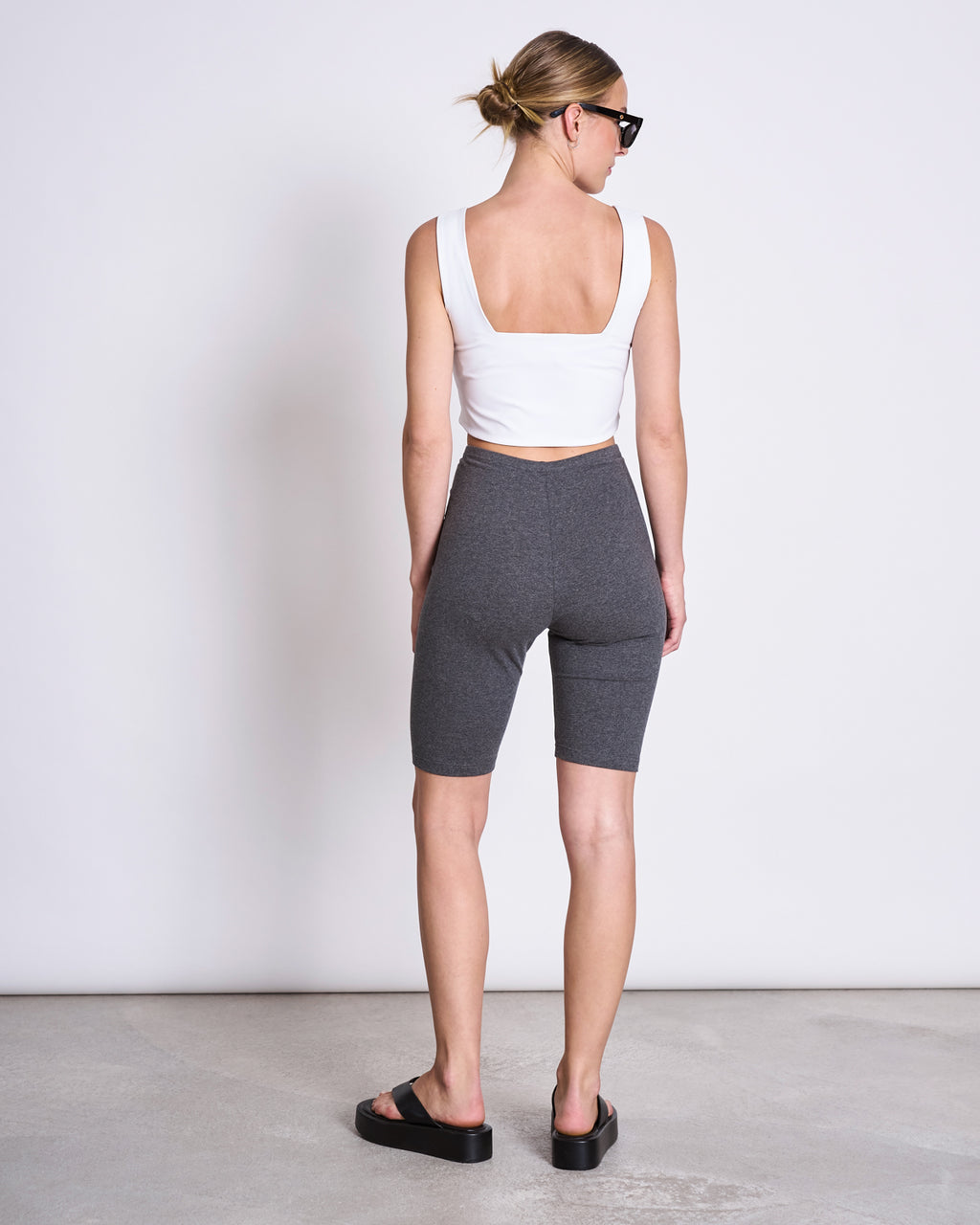 A woman with her hair in a low bun faces away from the camera, wearing jan-n-june BIKER SHORTS DARK GREY, a white sleeveless crop top, black slide sandals, and dark sunglasses. The background is plain.