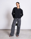 A woman poses in a minimalist setting wearing the jan-n-june BOXY LONGSLEEVE ANKIRA BLACK, gray high-waisted wide-leg jeans, black heeled sandals, and a silver necklace. She stands with one knee bent and hands behind her back.