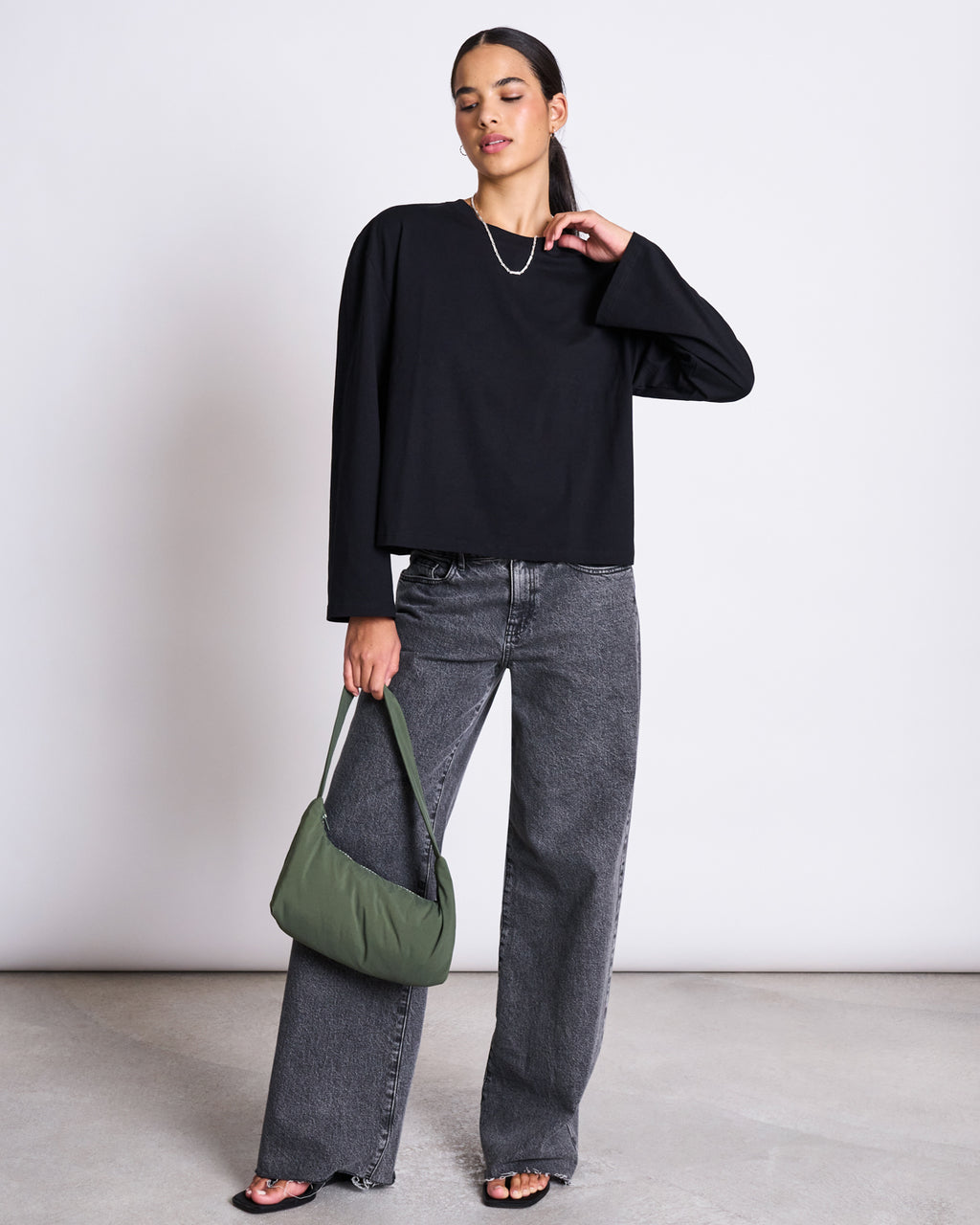 A woman stands against a plain background wearing the jan-n-june BOXY LONGSLEEVE ANKIRA BLACK top, wide-leg gray jeans, and black sandals. She holds a green handbag, wears a silver necklace, and has her hair pulled back.