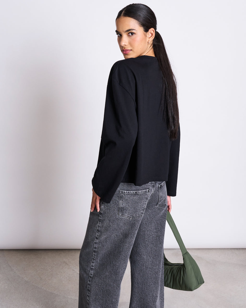 A woman with long dark hair in a ponytail wears the jan-n-june BOXY LONGSLEEVE ANKIRA BLACK top and gray wide-leg jeans, holding a small green handbag as she looks over her shoulder at the camera against a plain white background.