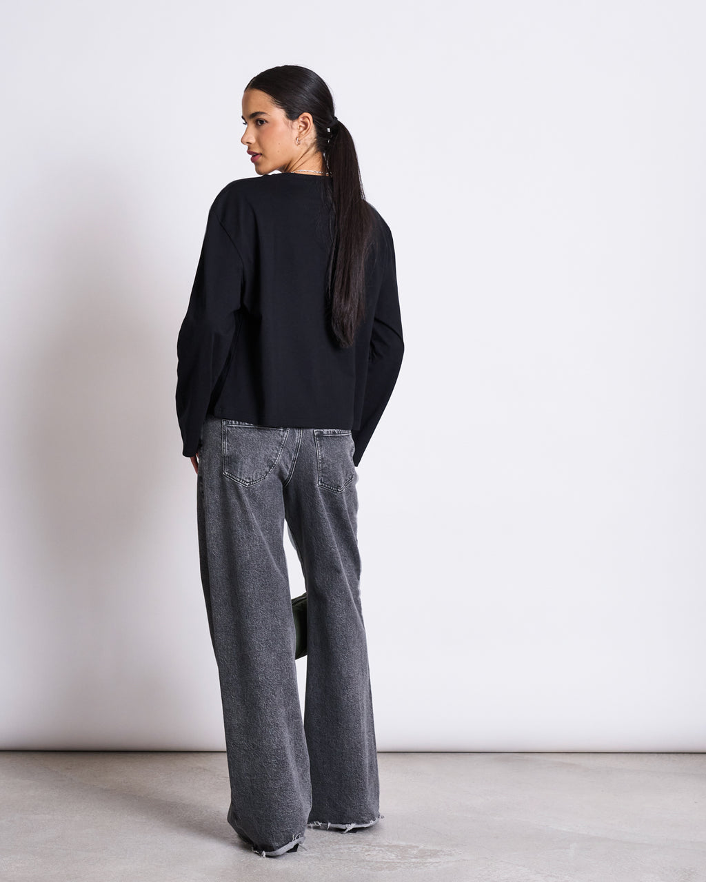 A woman with long dark hair in a low ponytail stands indoors, facing away. She wears the jan-n-june BOXY LONGSLEEVE ANKIRA BLACK and wide-leg gray jeans with frayed hems, set against a plain white background.