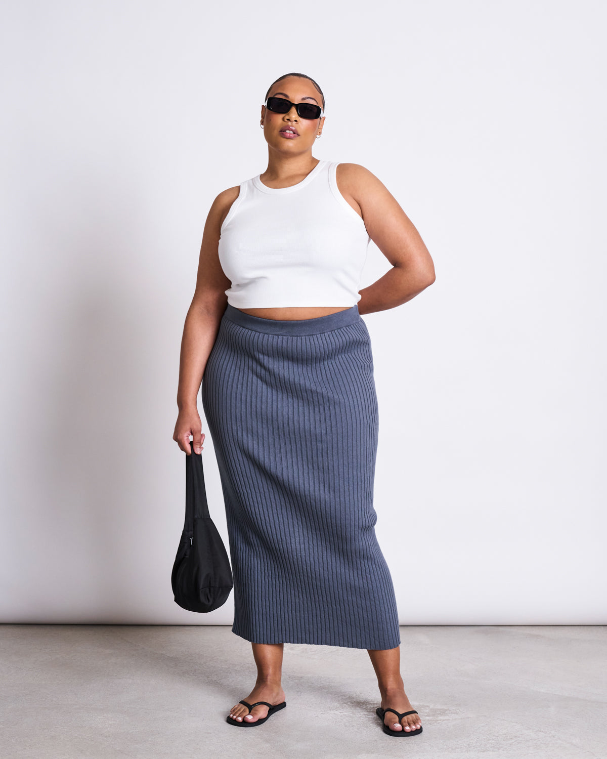 A woman in a white sleeveless crop top, black flip-flops, black sunglasses, and holding a small black bag wears the jan-n-june MIDI KNIT SKIRT EBBA TURBULENCE GOTS, standing against a plain white background.
