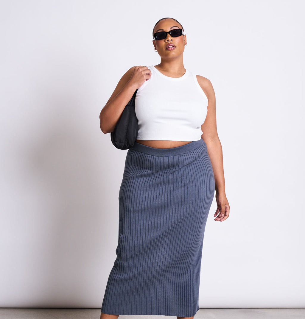 A person in the jan-n-june MIDI KNIT SKIRT EBBA TURBULENCE GOTS pairs it with a white sleeveless crop top, dark sunglasses, and a small black shoulder bag, standing against a plain white background.