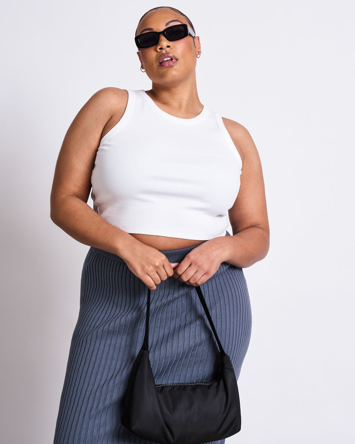 A woman holds a black handbag while wearing sunglasses, a white sleeveless crop top, and the jan-n-june MIDI KNIT SKIRT EBBA TURBULENCE GOTS in gray against a plain white background.