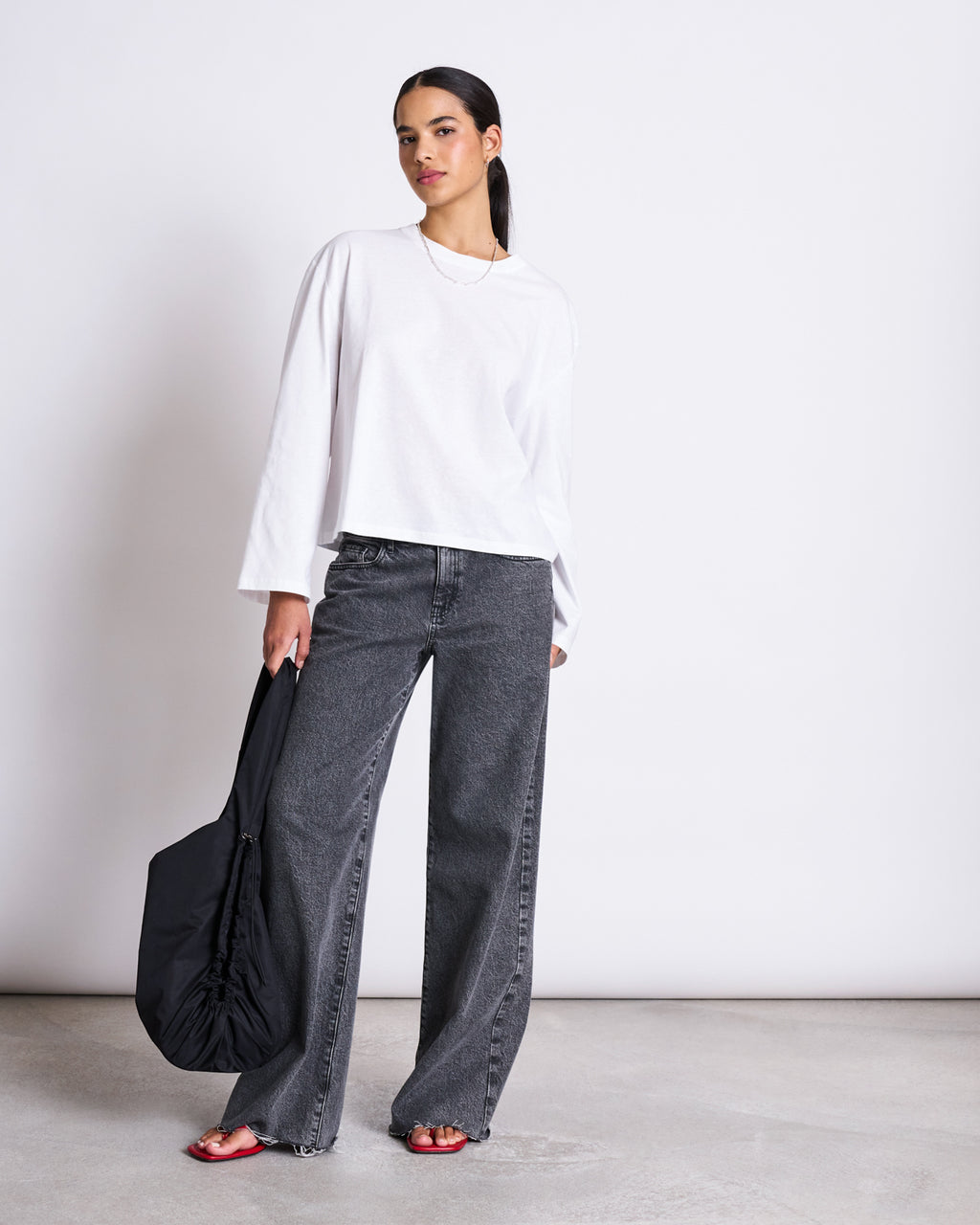 A woman in a BOXY LONGSLEEVE ANKIRA WHITE by jan-n-june, organic cotton, pairs it with wide-leg gray jeans, red sandals, and a black shoulder bag for an effortless oversized look against a plain background.