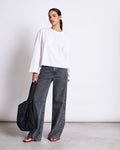A woman in a BOXY LONGSLEEVE ANKIRA WHITE by jan-n-june, organic cotton, pairs it with wide-leg gray jeans, red sandals, and a black shoulder bag for an effortless oversized look against a plain background.