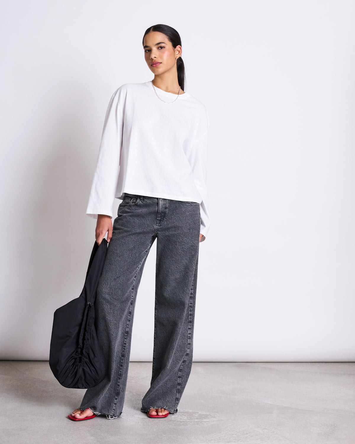 A woman in a BOXY LONGSLEEVE ANKIRA WHITE by jan-n-june, organic cotton, pairs it with wide-leg gray jeans, red sandals, and a black shoulder bag for an effortless oversized look against a plain background.