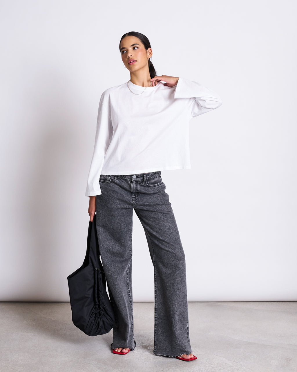 A woman stands against a white background wearing the jan-n-june BOXY LONGSLEEVE ANKIRA WHITE in organic cotton, wide-leg gray jeans, red sandals, and holding a large black tote. Her hair is tied back as she gazes slightly to the side.
