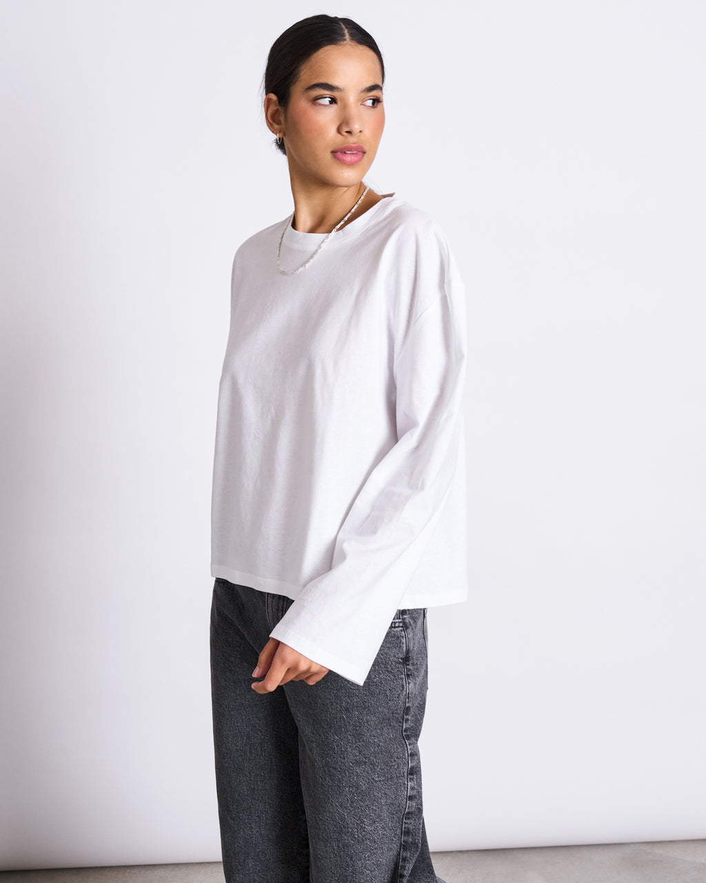 A person with dark hair tied back wears an oversized BOXY LONGSLEEVE ANKIRA WHITE by jan-n-june in organic cotton with faded black jeans, standing against a white background and looking to the side.